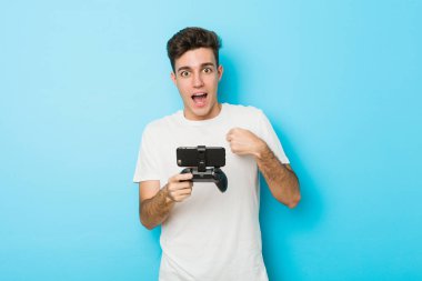 Kafkasyalı genç bir adam telefonuyla video oyunları oynuyor. Kendini işaret ederek, gülümseyerek şaşırdı..