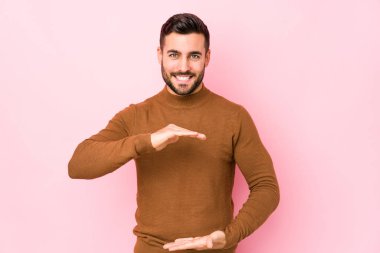 Young caucasian man against a pink background isolated holding something with both hands, product presentation.