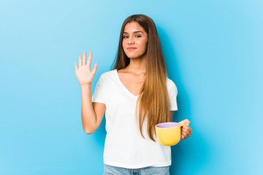 Young caucasian woman holding a coffee mug smiling cheerful showing number five with fingers.
