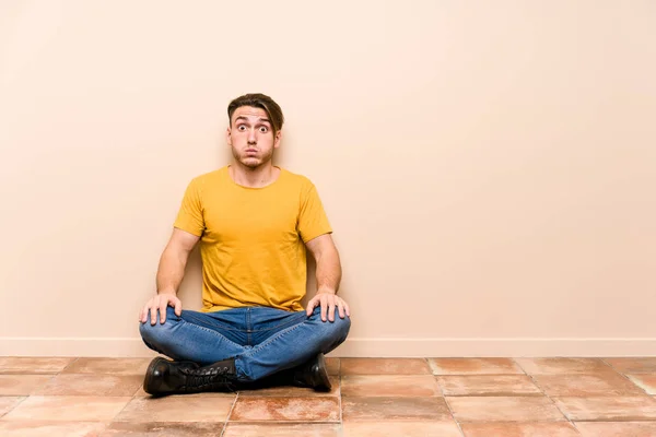 Young caucasian man sitting on the floor isolated blows cheeks, has tired expression. Facial expression concept.