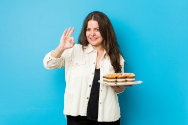 Genç, kıvrımlı bir kadın elinde kapkekleri neşeli ve kendinden emin bir şekilde tutuyor..