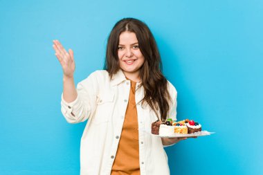 Genç, kıvrımlı bir kadın elinde tatlı bir pasta tutuyor. Hoş bir sürpriz alıyor, heyecanlı ve el kaldırıyor..