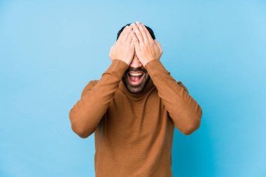 Mavi arka planda genç bir latin adam. Gözlerinizi ellerinizle kapayın. Gülümseyin ve bir sürpriz bekleyin..