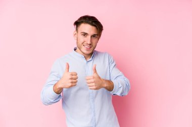 Pembe arka planda poz veren beyaz bir genç yalnız, iki parmağını da kaldırıyor, gülümsüyor ve kendinden emin..