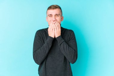 Mavi arka planda genç beyaz bir adam bir şeye gülüyor, elleriyle ağzını kapatıyor..