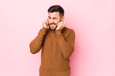 Pembe arka planda genç beyaz bir adam. Elleriyle kulaklarını kapatıyor..