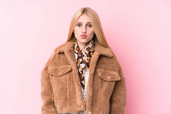Young blonde woman wearing a coat against a pink background blows cheeks, has tired expression. Facial expression concept.