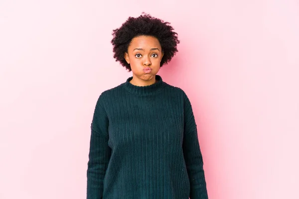 Middle aged african american woman against a pink background isolated blows cheeks, has tired expression. Facial expression concept.