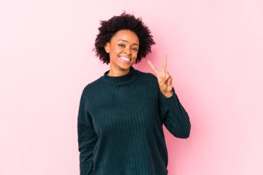 Orta yaşlı Afrikalı Amerikalı kadın pembe bir arka plana karşı izole edilmiş zafer işareti gösteriyor ve gülümsüyor..