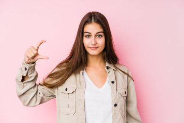 Pembe arka planda poz veren beyaz bir kadın. Elinde parmaklarıyla küçük bir şey tutuyor, gülümsüyor ve kendinden emin..