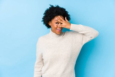 Orta yaşlı Afrikalı Amerikalı bir kadın mavi bir geçmişe karşı kameraya parmaklarıyla göz kırpıyor, yüzünü kapatmaktan utanıyor..