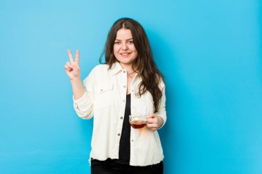 İki numarayı parmaklarıyla gösteren bir çay fincanı tutan kıvrımlı genç kadın..