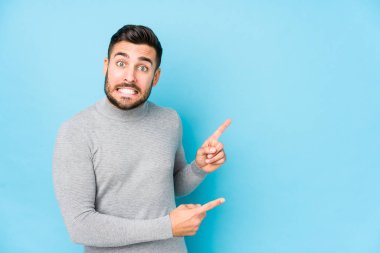 Kafkasyalı genç adam mavi arka planda, izole edilmiş, şok olmuş, işaret parmaklarıyla bir kopya alanı işaret ediyor..