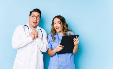 Mavi arka planda poz veren genç doktor çift şans için dua ediyor, hayret içinde ve ağzı açık bir şekilde öne bakıyor..