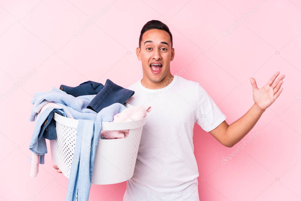 Young latin man picking up dirty clothes isolated receiving a pleasant surprise, excited and raising hands.