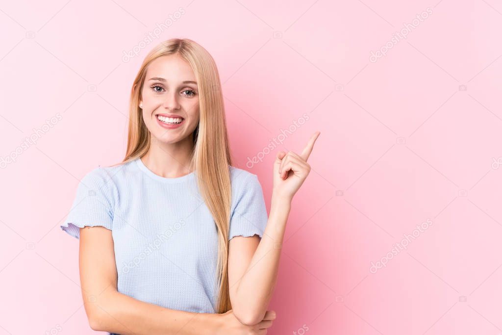 Young blonde woman on pink background smiling cheerfully pointing with forefinger away.
