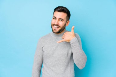 Mavi arka planda genç beyaz bir adam izole edilmiş, parmakları olan bir cep telefonu hareketi gösteriyor..