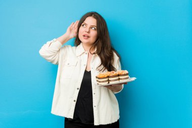 Genç, kıvrımlı bir kadın elinde keklerle bir dedikoduyu dinlemeye çalışıyor..