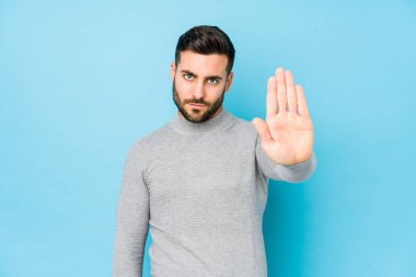 Kafkasyalı genç bir adam mavi arka planda izole edilmiş, elini uzatmış, dur işareti gösteriyor, bu da seni engelliyor..
