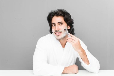 Gri bir arka planda sakalını kesen genç beyaz bir adam.