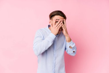 Pembe arka planda poz veren beyaz bir genç parmaklarıyla korkmuş ve gergin bir şekilde göz kırpıyor..