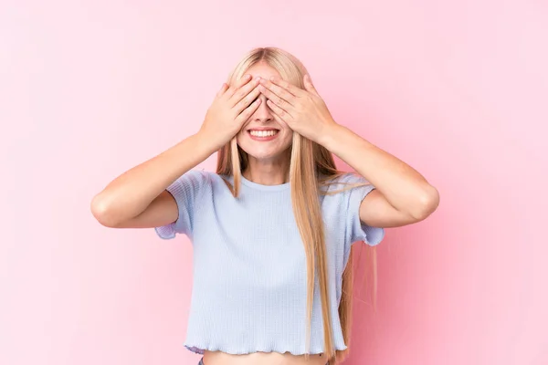 Young blonde woman on pink background covers eyes with hands, smiles broadly waiting for a surprise.