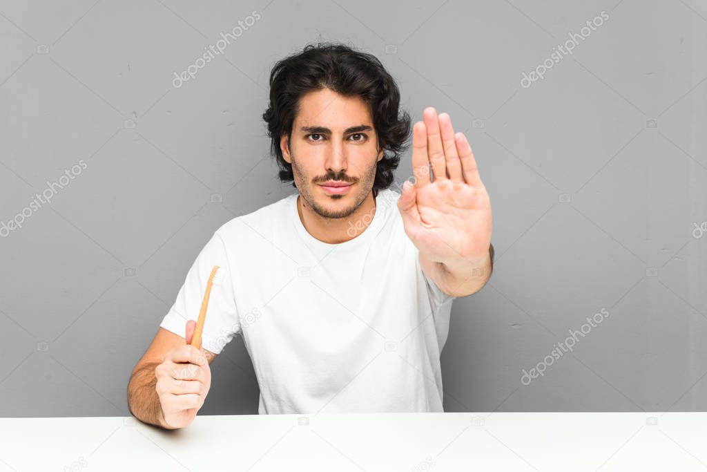 Young man holding a toothbrush standing with outstretched hand showing stop sign, preventing you.