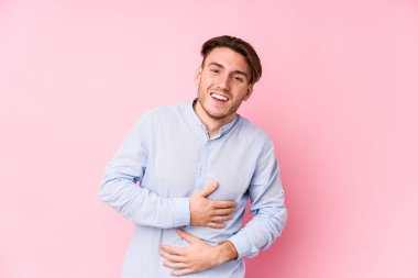 Pembe arka planda poz veren beyaz bir genç mutlu bir şekilde gülüyor ve ellerini karnının üstünde tutarak eğleniyor..