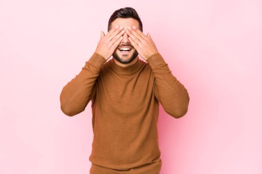 Pembe arka planda genç beyaz bir adam izole edilmiş gözleri ellerle kaplıyor, geniş bir gülümsemeyle bir sürpriz bekliyor..