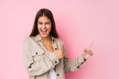 Young caucasian woman posing in a pink background pointing with forefingers to a copy space, expressing excitement and desire.