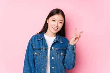 Pembe arka planda poz veren genç Çinli kadın neşeli ve tasasız bir şekilde parmaklarıyla barış sembolü gösteriyor..