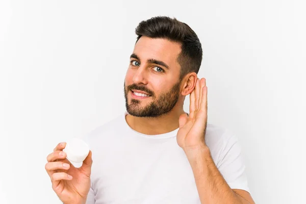 Joven Hombre Caucásico Sosteniendo Una Crema Hidratante Aislado Tratando Escuchar —  Fotos de Stock