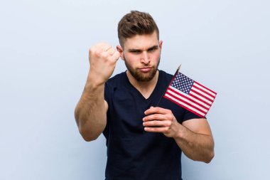 Amerika Birleşik Devletleri bayrağını tutan genç beyaz bir adam kameraya yumruğunu gösteriyor, agresif bir yüz ifadesi..
