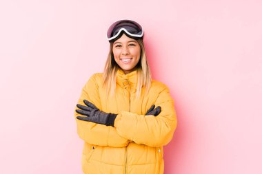 Pembe arka planda kayak kıyafeti giyen genç beyaz bir kadın kendini güvende hissediyor, kararlılıkla kollarını kavuşturuyor..