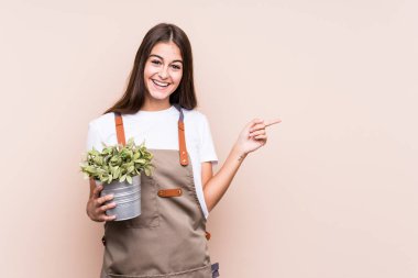 Young gardener caucasian woman holding a plant isolatedsmiling and pointing aside, showing something at blank space.