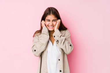 Young caucasian woman posing in a pink background covering ears with hands.