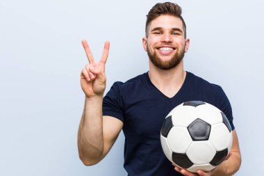 Genç beyaz bir adam elinde futbol topu tutuyor. İki numarayı parmaklarıyla gösteriyor..