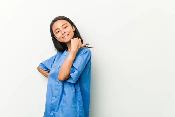 Young asian nurse woman points with thumb finger away, laughing and carefree.