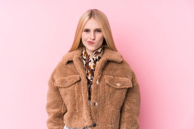 Pembe arka planda palto giyen, hoşnutsuzca kaşlarını çatan, kollarını kavuşturan genç sarışın kadın..