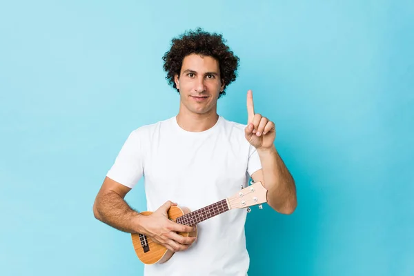 Young caucasian curly man playing ukelele showing number one with finger.