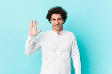 Genç, kıvırcık, olgun bir adam zarif bir gömlek giyiyor. Gülümsüyor. Beş numarayı parmaklarıyla gösteriyor..