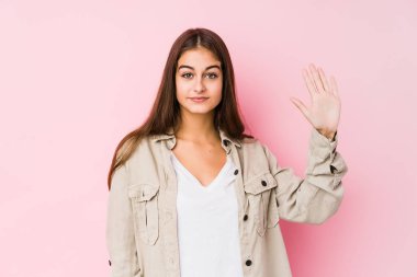Pembe arka planda poz veren beyaz bir kadın gülümseyerek beş numarayı parmaklarıyla gösteriyor..