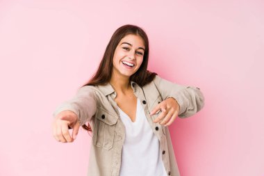 Young caucasian woman posing in a pink background cheerful smiles pointing to front.