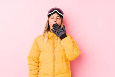 Pembe arka planda kayak kıyafeti giyen genç beyaz kadın esniyor. Ağzı eliyle kapatan yorgun bir jest sergiliyor..