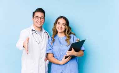 Mavi arka planda poz veren genç doktor çift kameraya el uzatarak selam veriyor..