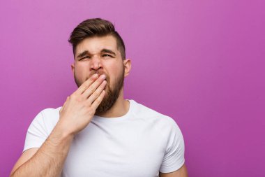 Young handsome caucasian man yawning showing a tired gesture covering mouth with him hand.
