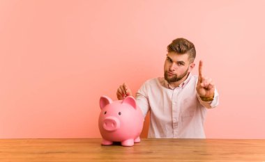 Young caucasian man sitting with a piggy bank showing number one with finger.