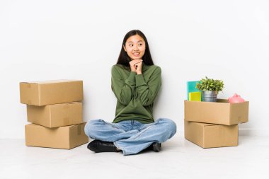 Young chinese woman moving to a new home keeps hands under chin, is looking happily aside. clipart