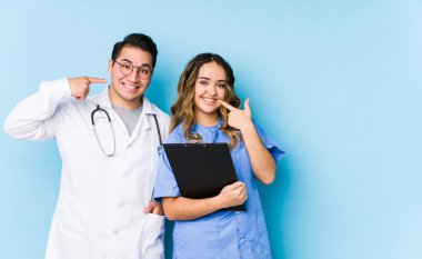 Mavi arka planda poz veren genç doktor çift, izole edilmiş gülümsemeler, parmaklarını ağıza doğrultuyor..