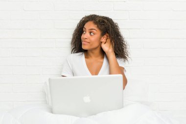 Young african american woman sitting on the bed with laptop trying to listening a gossip.